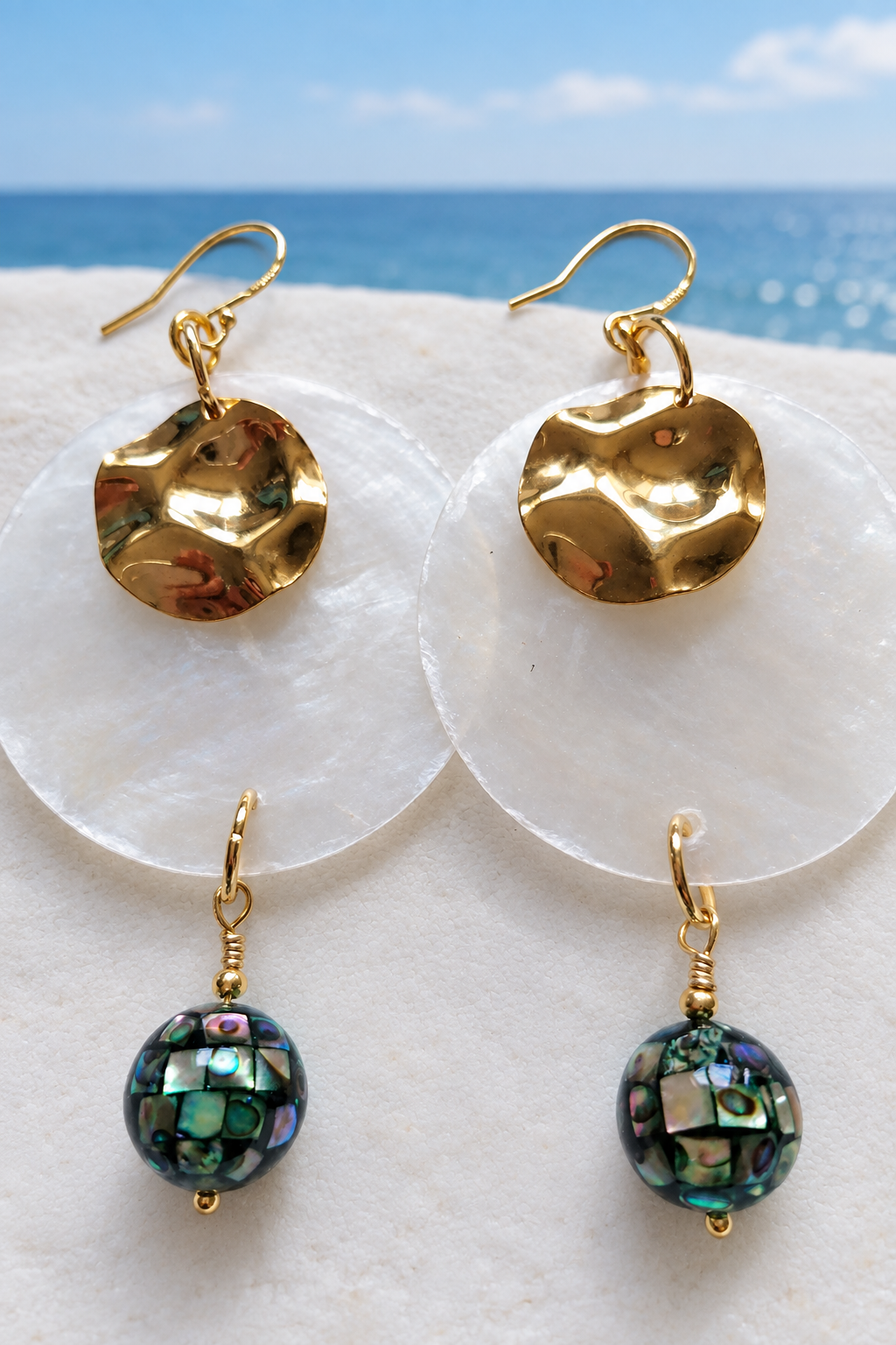 Gold hammered disc earrings with translucent white shell discs and abalone shell mosaic ball drops, laid flat on a white surface with a coastal blue sky horizon