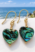 Abalone shell heart earrings on 24ct gold vermeil ear hooks, laid on a white surface with a coastal blue sky horizon