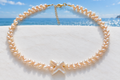 Peach freshwater pearl necklace with cross-shaped baroque pearl centrepiece and gold lobster clasp on a white surface with coastal sea view