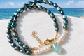 Peacock baroque pearl and white pearl necklace with aqua teardrop gemstone and gold lobster clasp on a white surface with coastal sea view