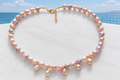 Pink and peach baroque freshwater pearl necklace with pearl drops and gold lobster clasp on a white surface with coastal sea view