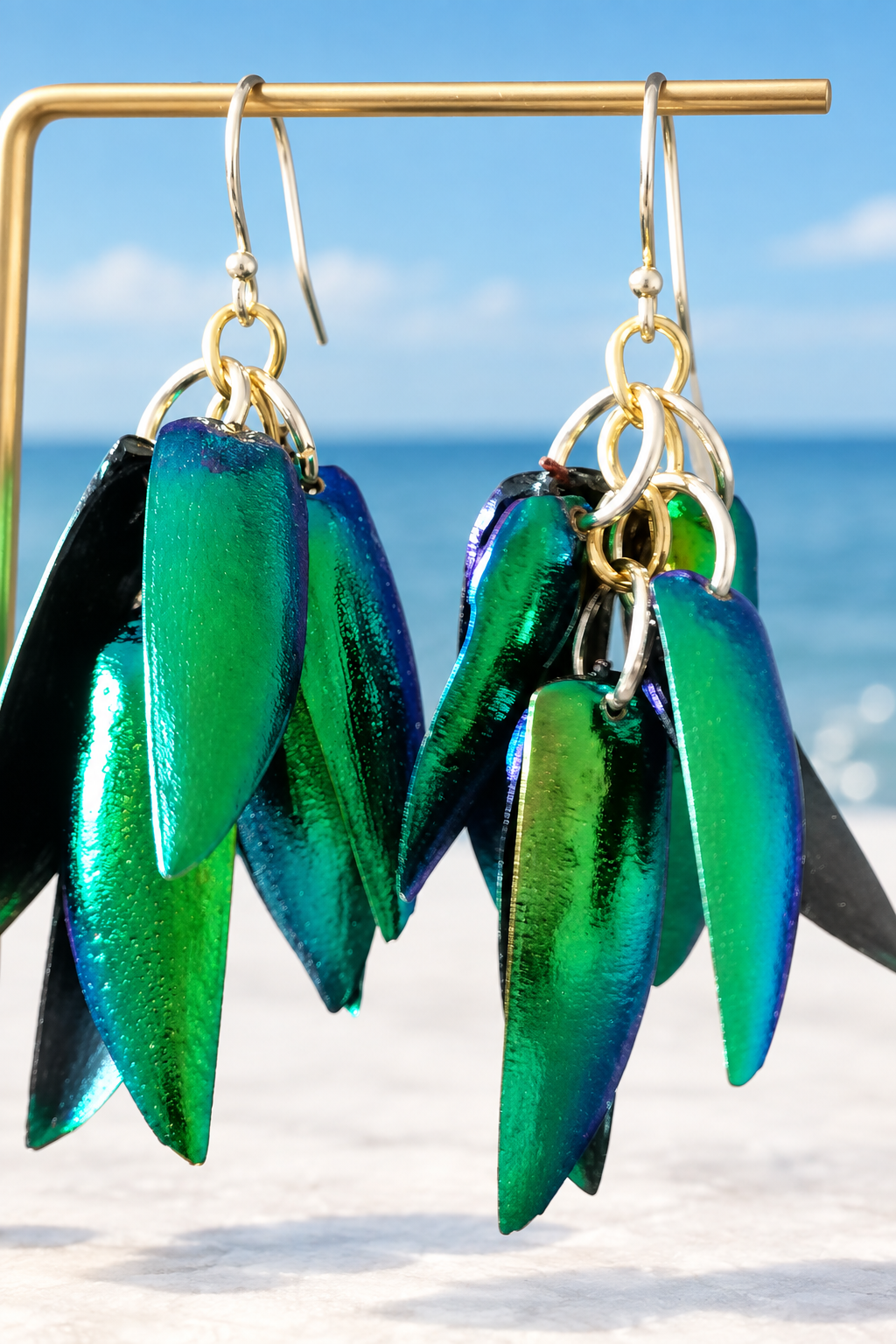 Iridescent green jewel beetle wing cluster earrings hanging on a gold stand with a coastal blue sky and sea background